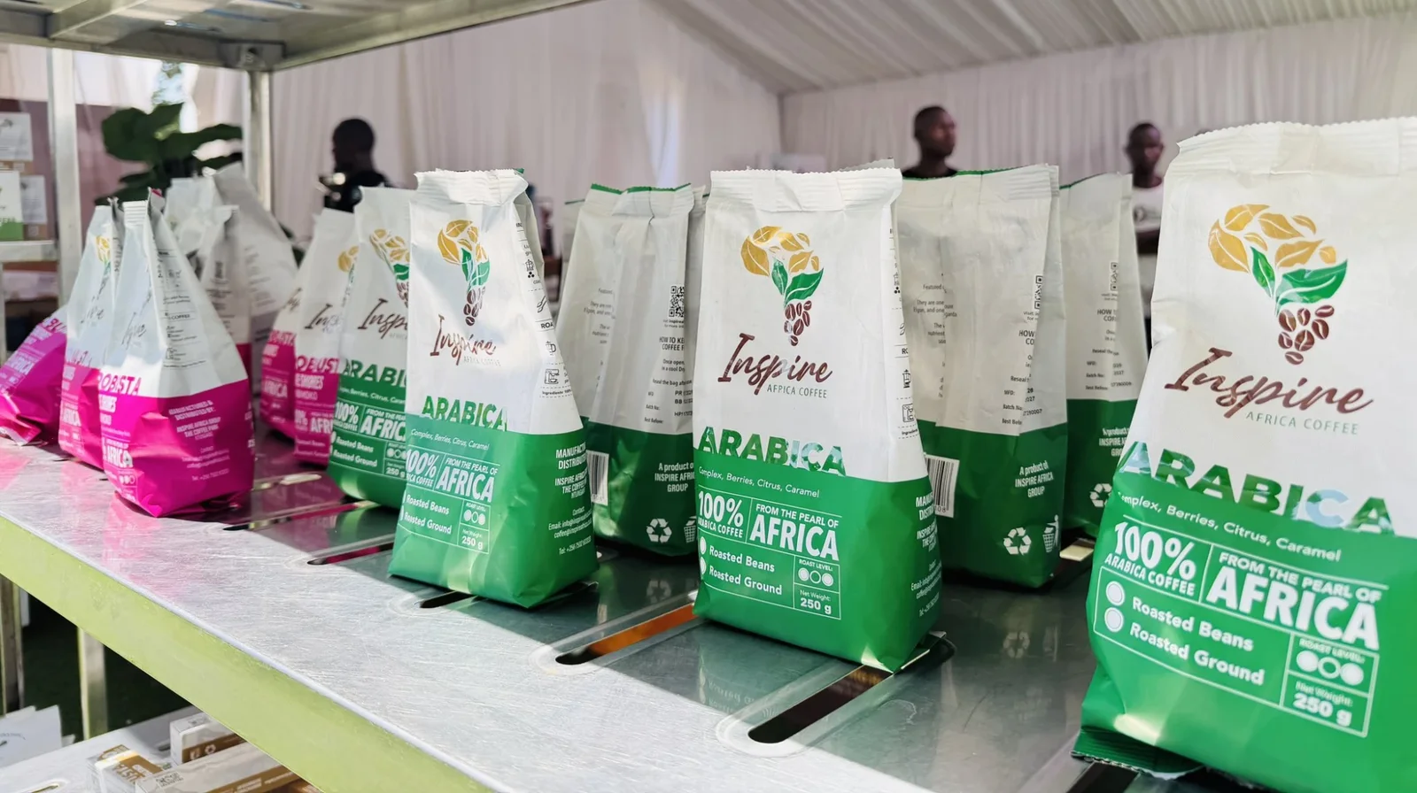 A row of 250g Inspire Africa Coffee Arabica packs on a stand shelf, green-and-white bags reading 100% Arabica Coffee, From the Pearl of Africa, with pink Robusta packs behind them