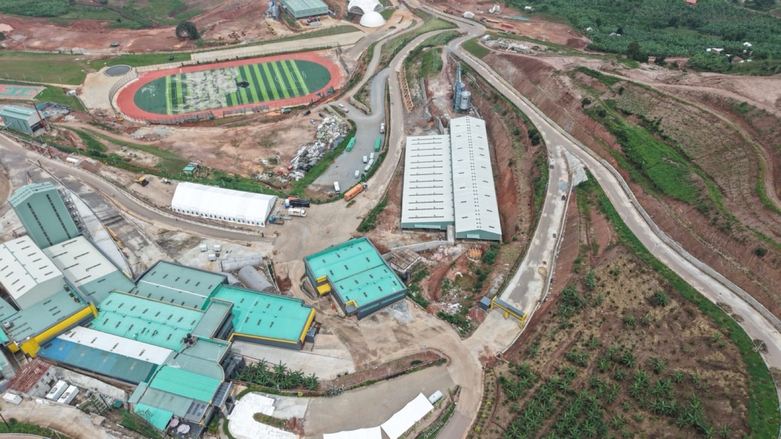The Africa Coffee Park processing complex seen from directly above: mill and roastery buildings, long warehouse sheds, storage silos, the athletics track, and haul roads cut into the hillside at Rwashameire