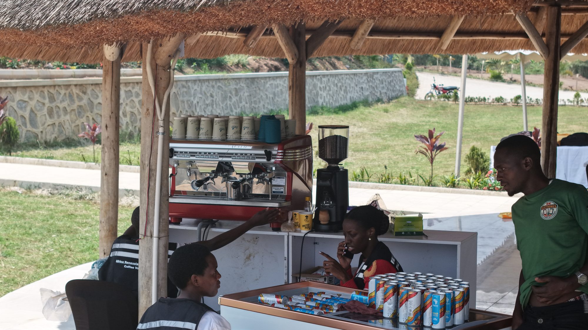 The Inspire coffee bar under the thatched lakeside pavilion at Africa Coffee Park, espresso machine running and Inspire Cold Brew cans stacked on the counter.