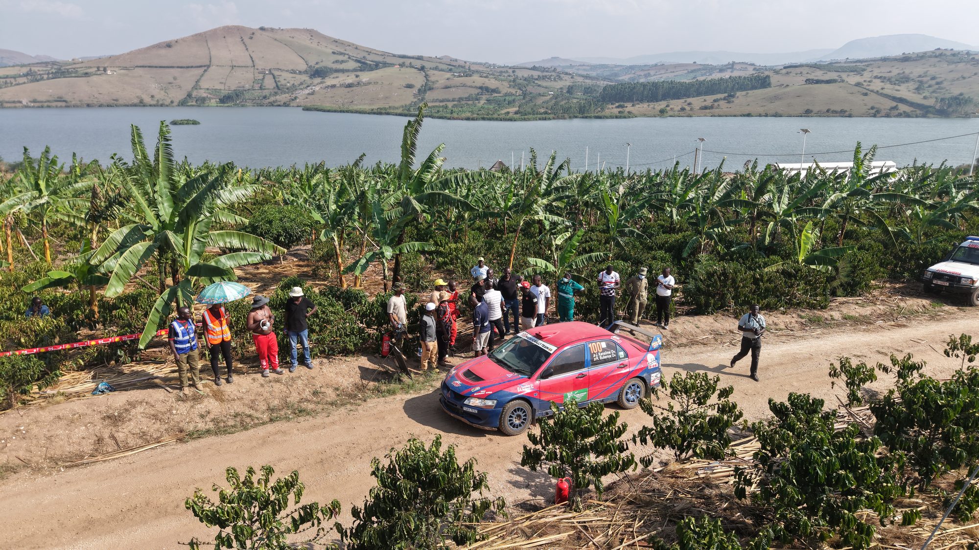 Aerial view of a rally car on the estate stage at Africa Coffee Park, spectators lining a dirt track that runs between coffee and banana with Lake Nyabihoko behind.