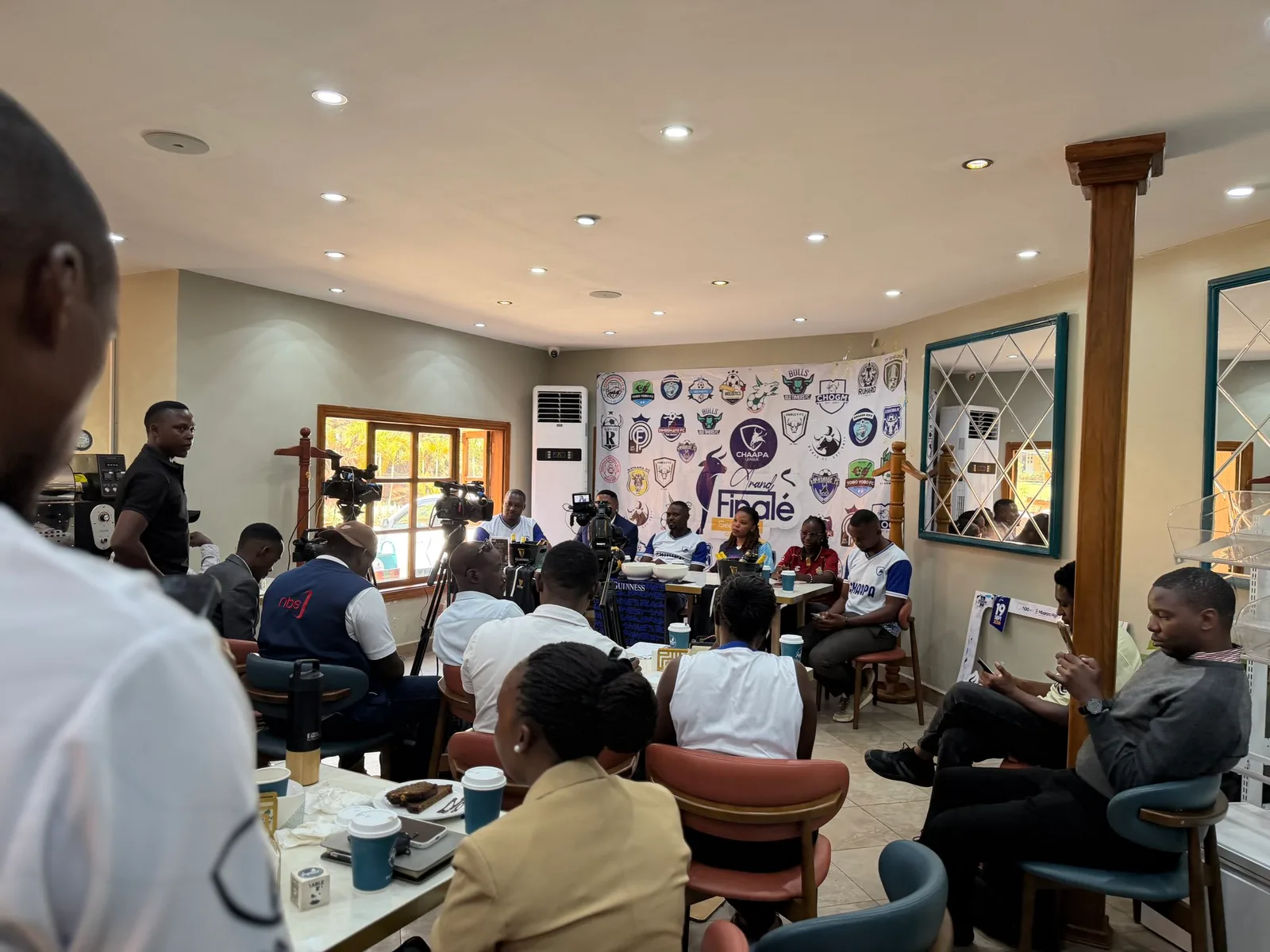 The press room at Inspire Café: a table of speakers in front of the Chaapa League backdrop, with camera operators and guests seated around the café.