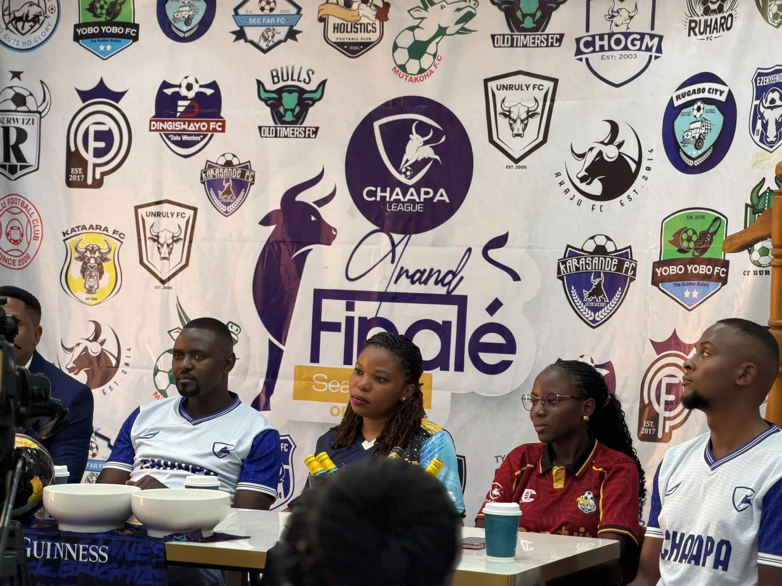 Four people seated at a table in front of a large Chaapa League Grand Finalé backdrop covered in club crests, two of them in club jerseys, with coffee cups on the table.