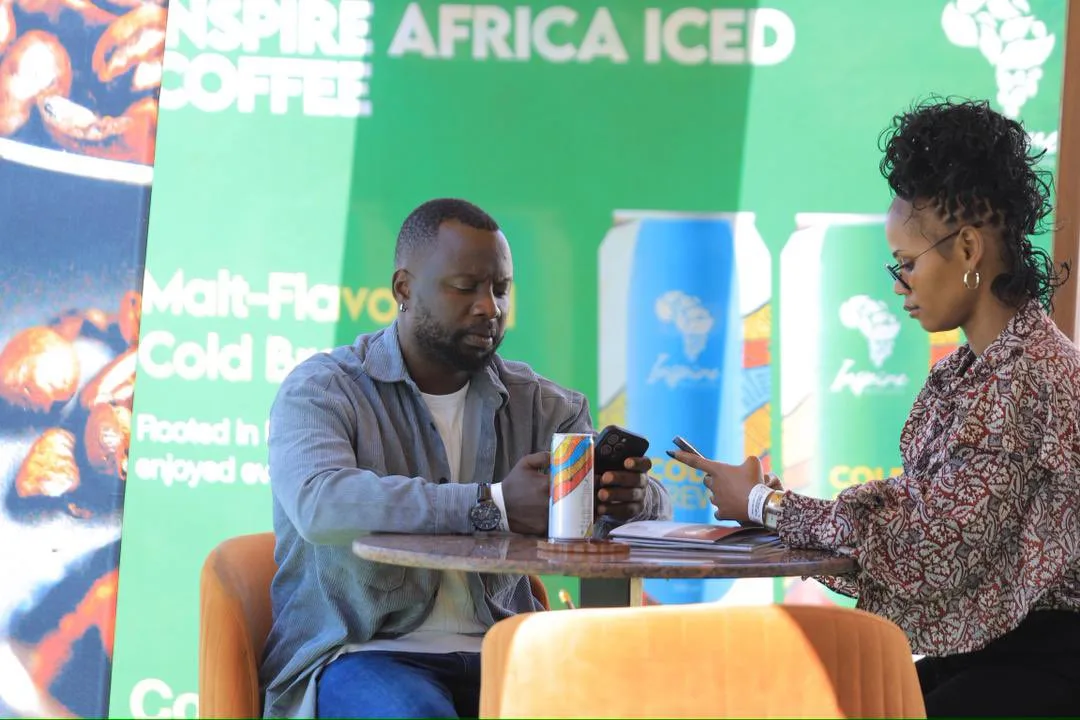 Two people at a small table in front of a green Inspire Africa Iced Coffee backdrop, one holding a can of cold brew, with more cans pictured on the banner behind them.