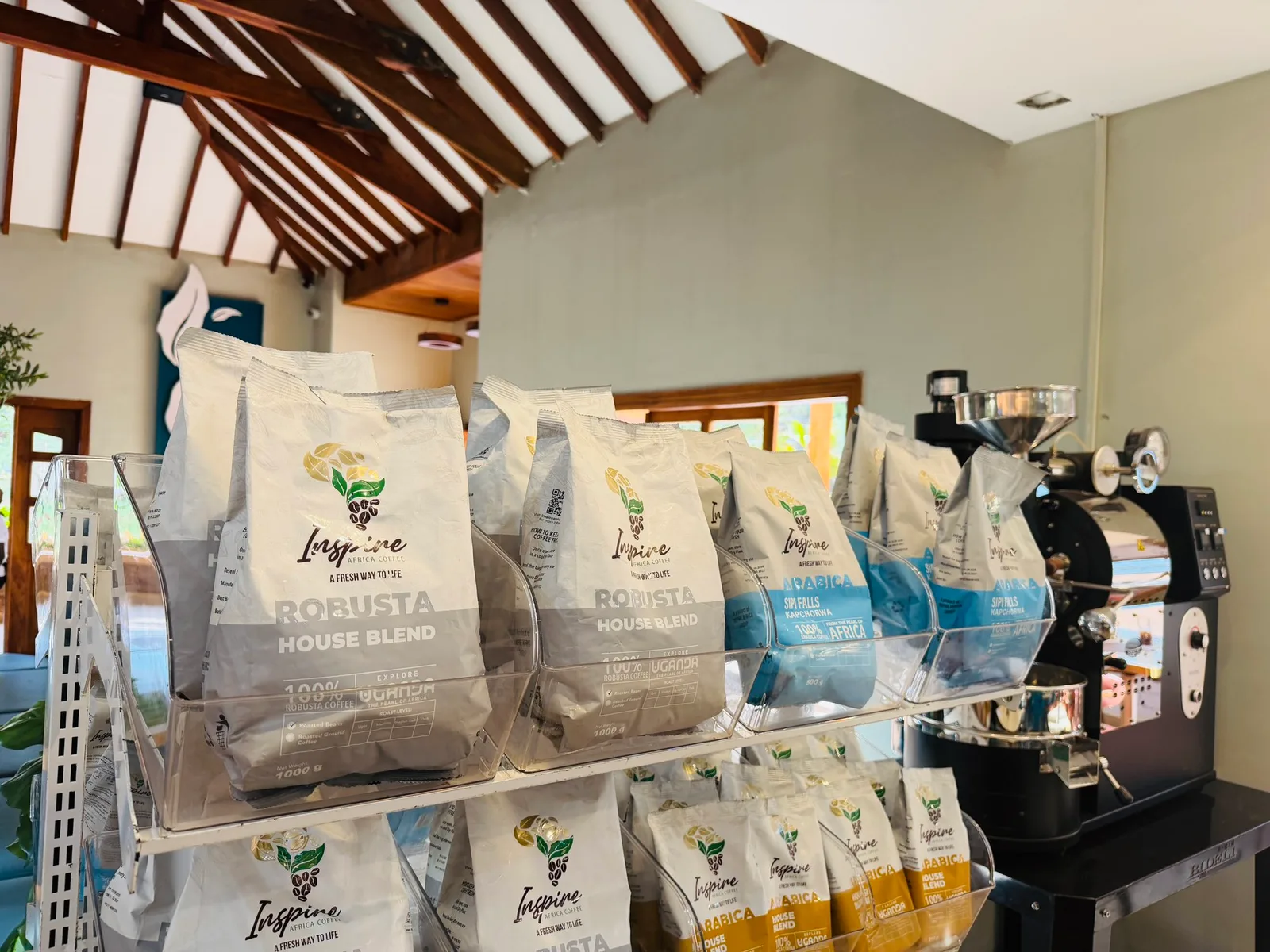 Bags of Inspire Robusta House Blend and other roasted coffee on clear display racks inside Inspire Café, with a black machine on a stand beside them.