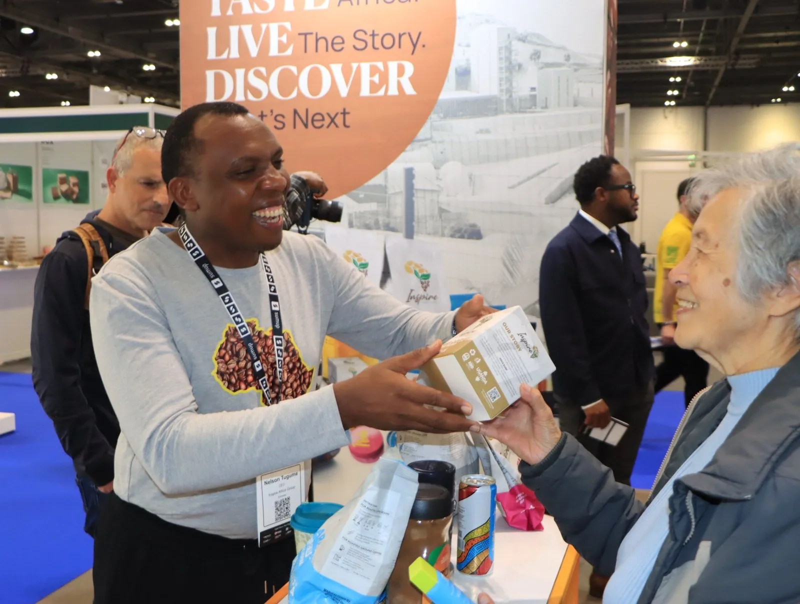 Nelson Tugume, whose badge reads CEO, Inspire Africa Group, handing a box of Inspire coffee to a smiling visitor at ExCeL London.