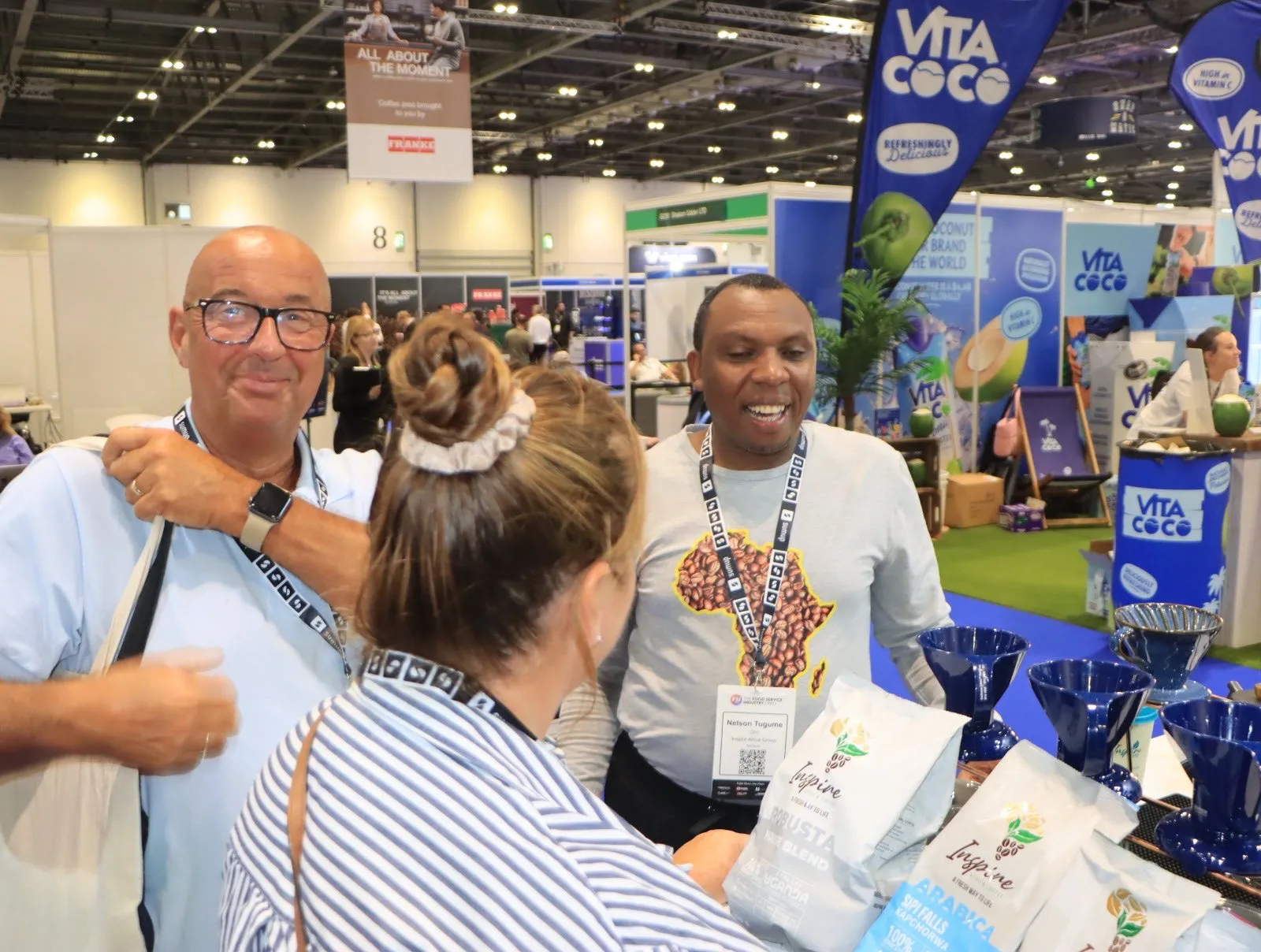 Visitors and a member of the Africa Coffee Park team smiling at a stand at ExCeL London, with bags of Inspire coffee and blue pour-over drippers on the counter.