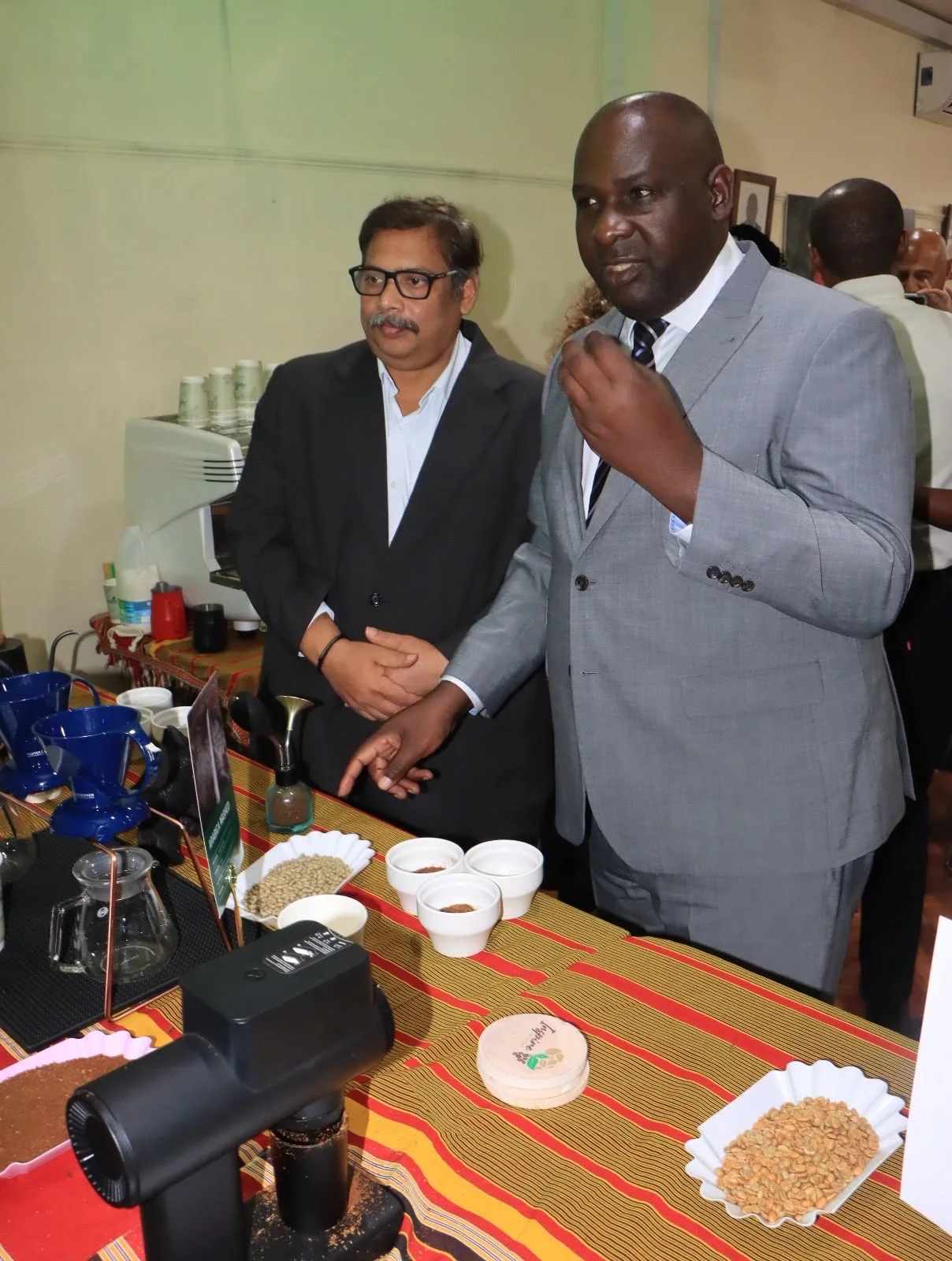 Two men in suits talking beside a tasting table set with white cups, a grinder and bowls of beans at the Uganda House showcase.