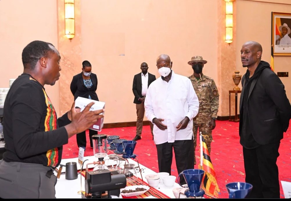 Dr. Nelson Tugume holding out a coffee pack across a brew bar of pour-over drippers, a grinder and cups, with President Museveni in a white shirt and Tyrese Gibson in black looking on, Uganda flags on the table