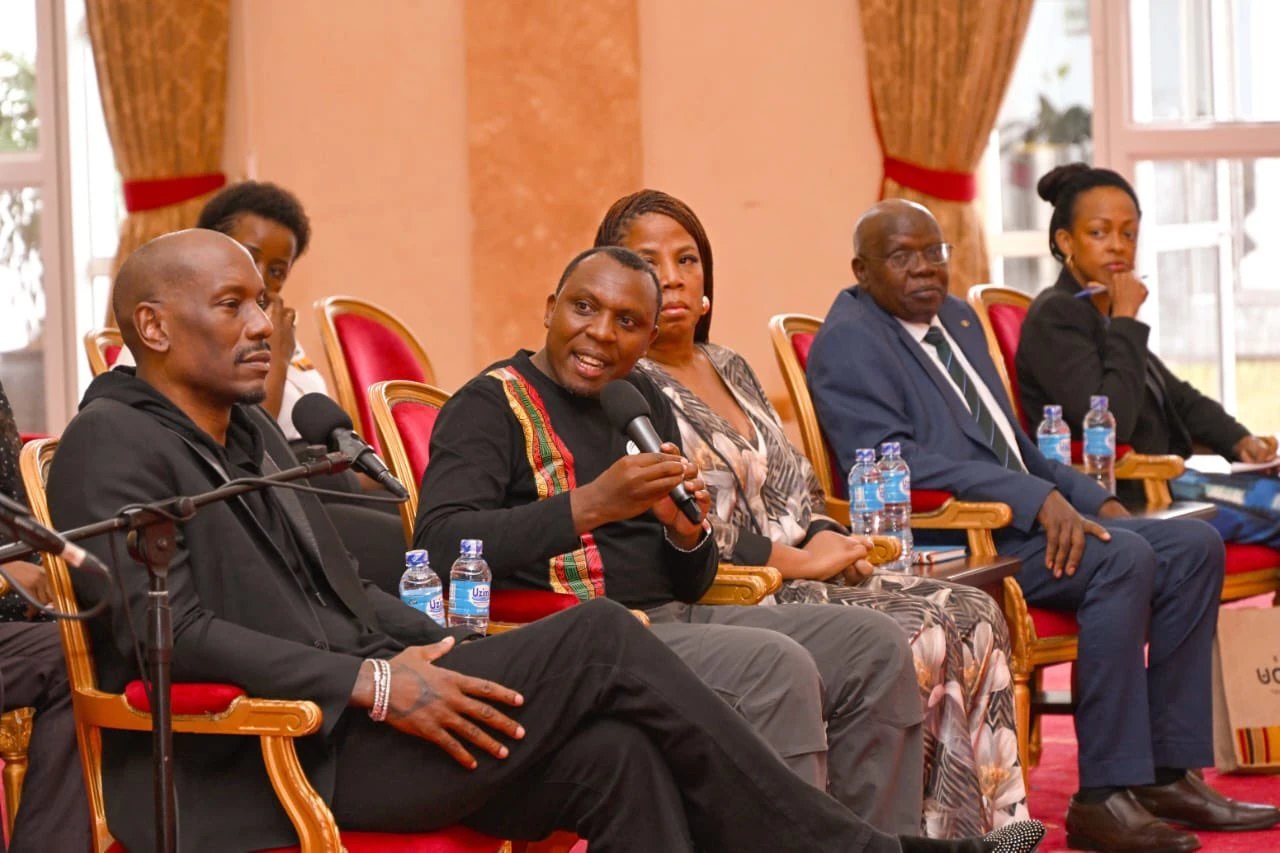 Tyrese Gibson seated on the left listening as Dr. Nelson Tugume speaks into a microphone on a panel of guests in gilt chairs at State House, Entebbe.