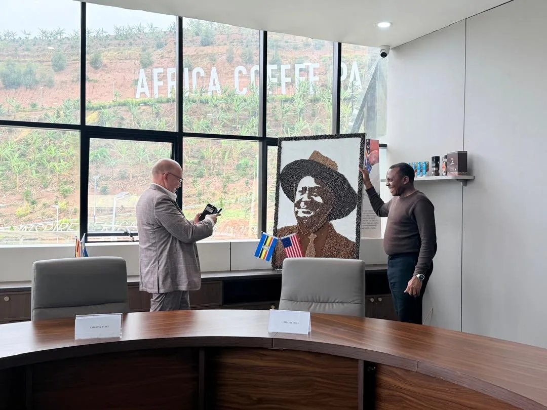 Two men in a meeting room at the park, one holding a phone and the other reaching up to a large portrait made of coffee beans, with an American flag and Embassy place cards on the table and the hillside lettering AFRICA COFFEE PARK visible through the window.