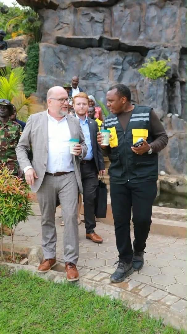 Two men in conversation holding teal paper coffee cups on a paved path beside a rock wall, with security personnel and other visitors behind them.