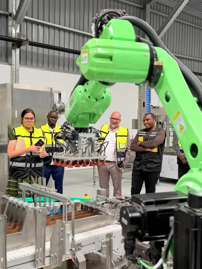 Four people in high-visibility vests watching a large green robotic arm positioned over a packaging line inside the plant.