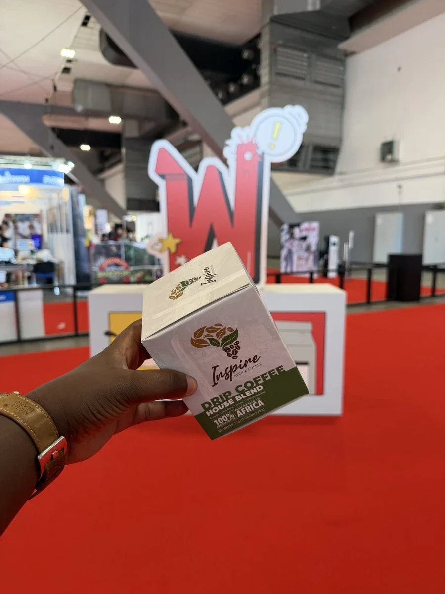A hand holding an Inspire Africa Drip Coffee House Blend box in the exhibition hall at World of Coffee Brussels