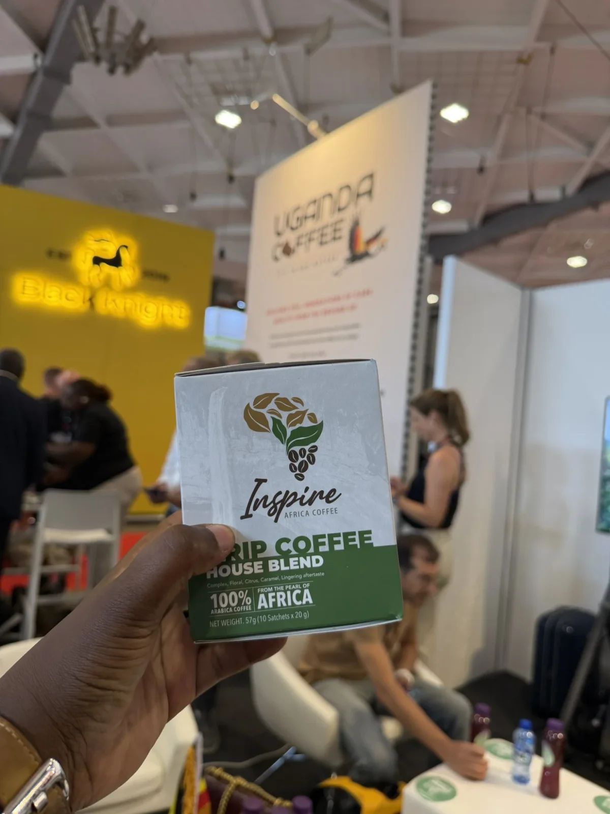 A hand holding an Inspire Africa Drip Coffee House Blend box in front of the Uganda Coffee stand at World of Coffee Brussels